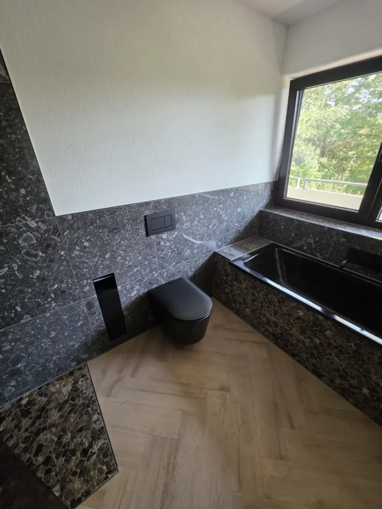 Schwarzes Wand-WC und Badewanne mit Steinoptikfliesen und Boden in Holzoptik.