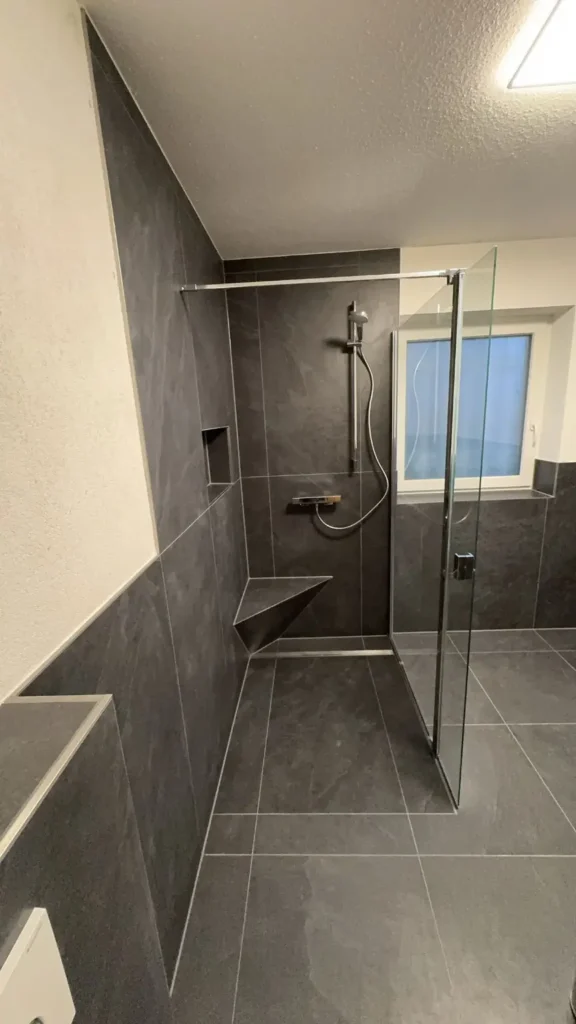 Geflieste Dusche mit Glasabtrennung, grauen Wandfliesen, Wandnische und dreieckiger Sitzbank.